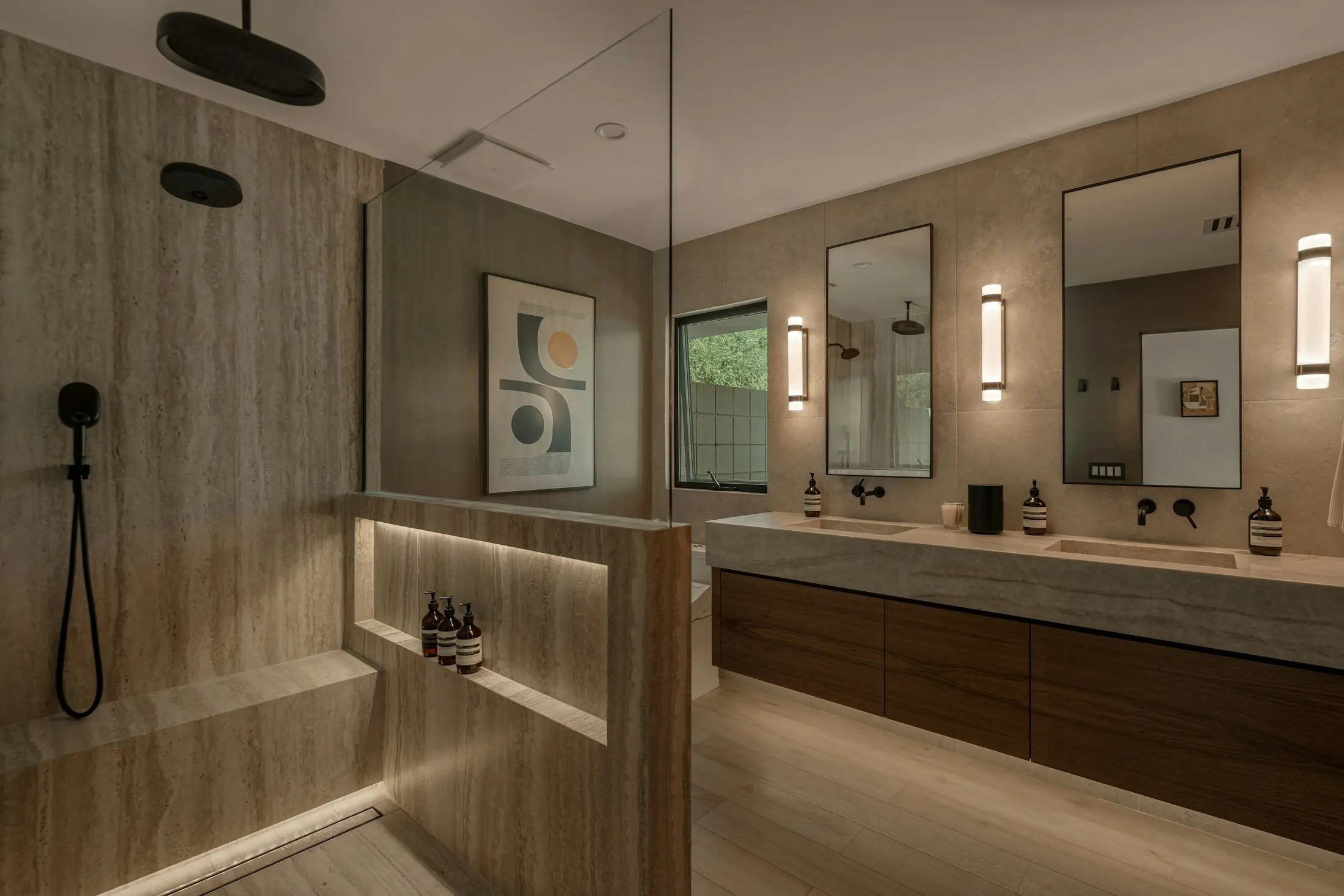 A finished primary bathroom: travertine-look porcelain walls, a walk-in shower with a lit bench and a double vanity